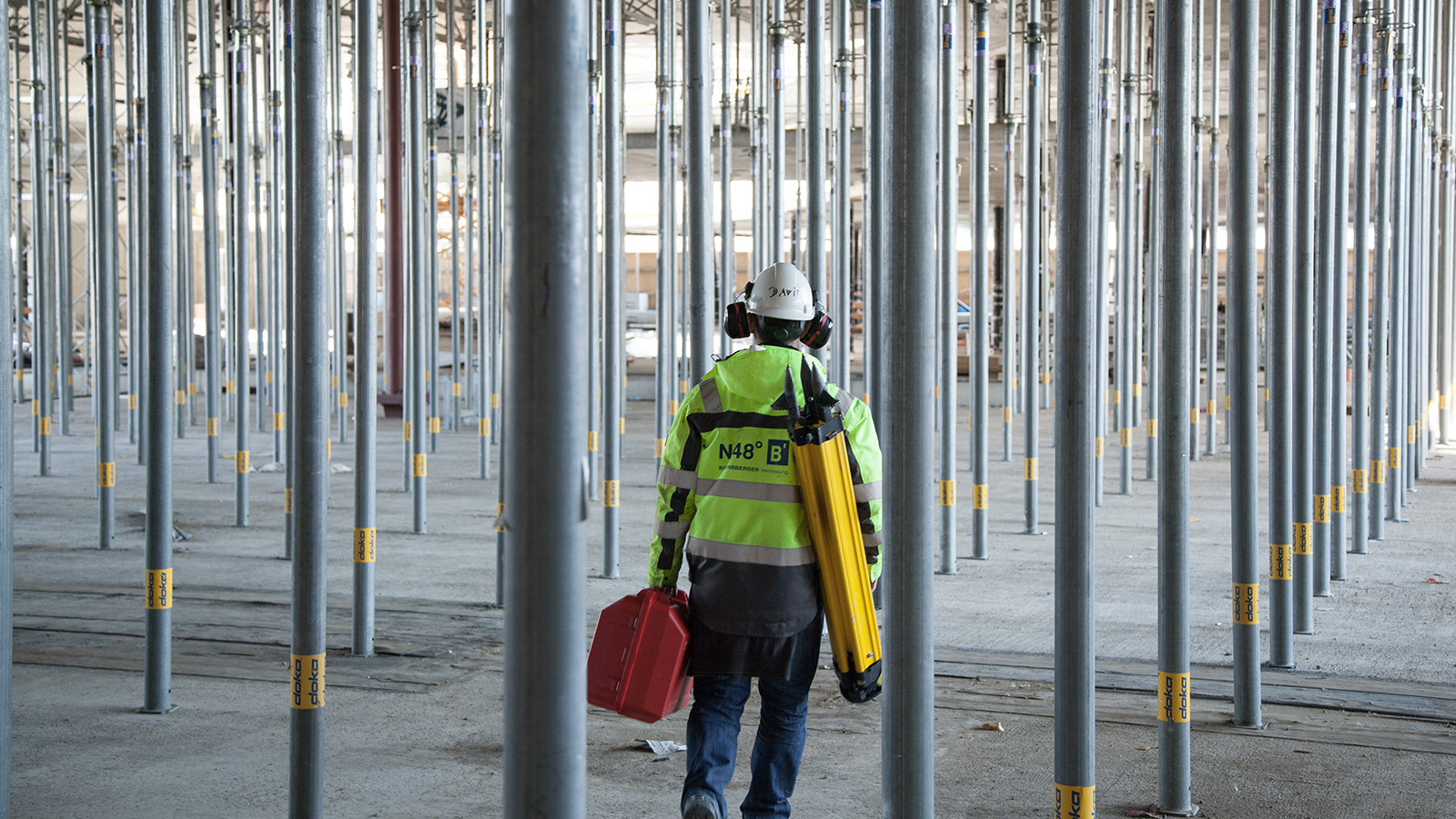 Ein Mitarbeiter von Bogensberger mit Stativ und Koffer auf einer Baustelle inmitten von Stützpfeilern