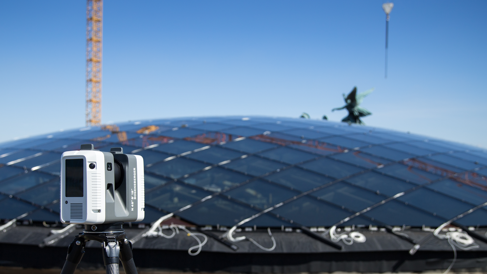 Ein Laserscanner RTC auf einem Stativ vor dem Dach des Parlaments mit Glasmodulen; blauer Himmel, Kran im Hintergrund.
