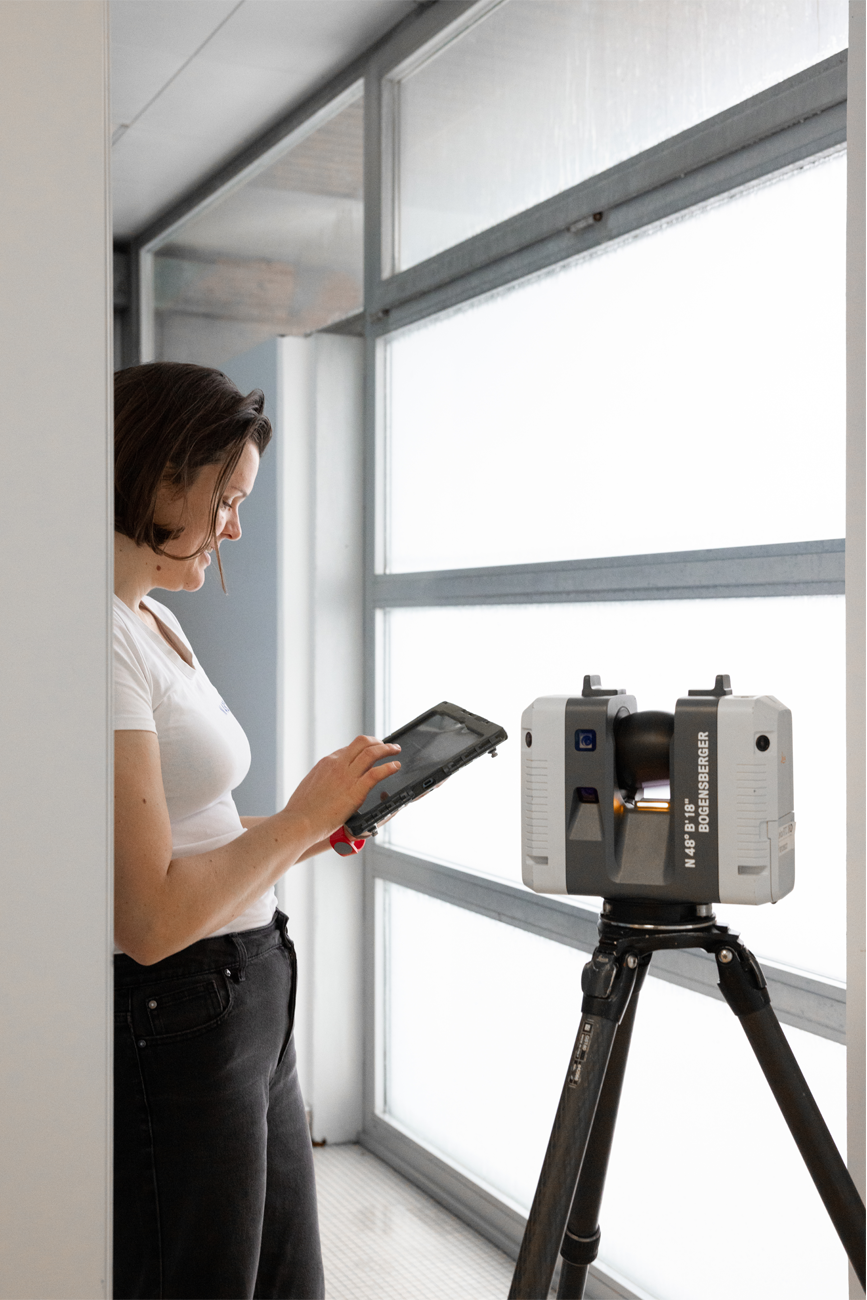 Eine Person steht am Fenster und bedient ein Tablet; davor ein Totalstation-Vermessungsgerät auf einem Stativ.