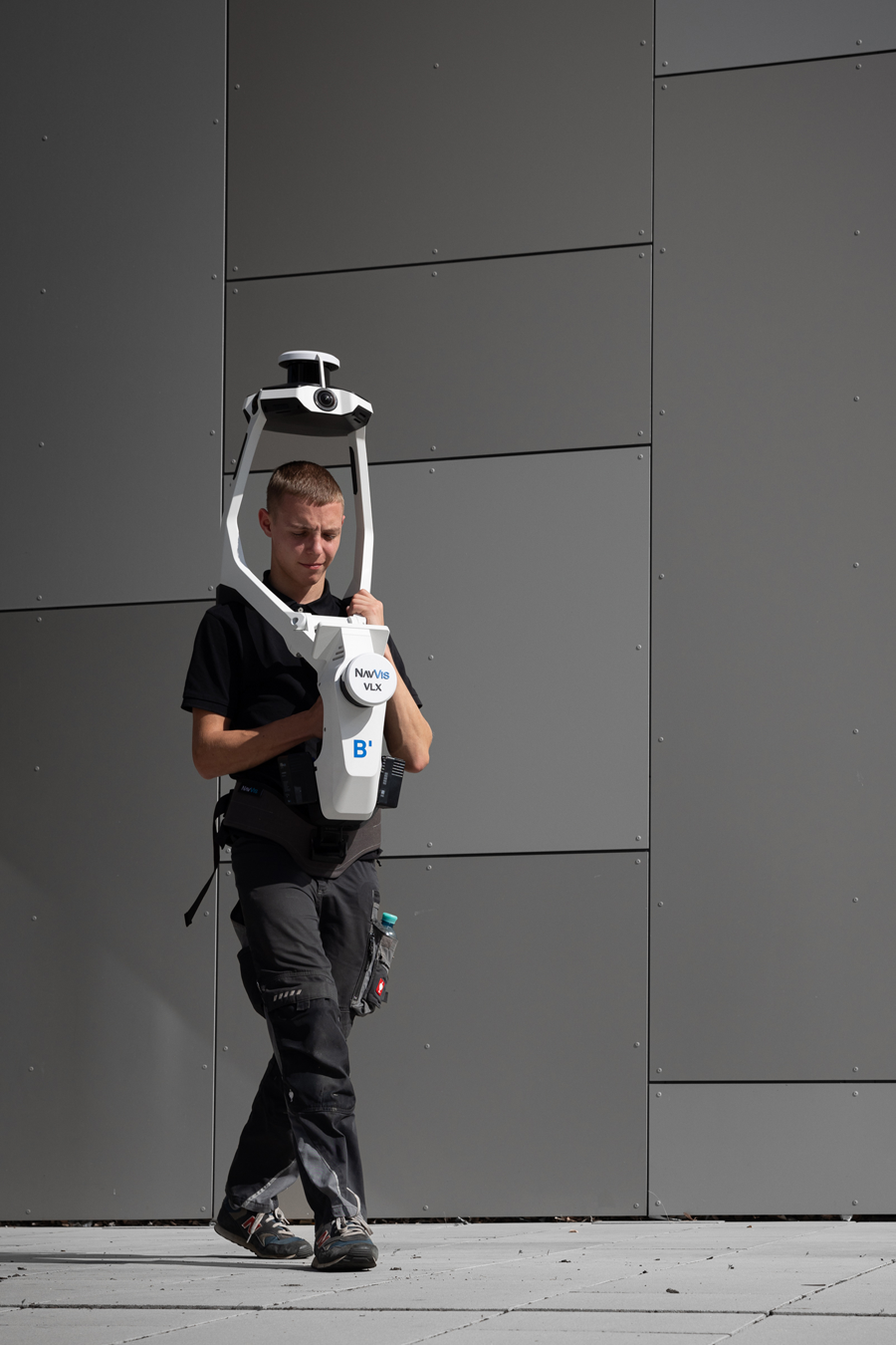 Eine Person steht vor einer grauen Metallwand und trägt ein großes, weißes NavVis VLX-Vermessungsgerät um den Oberkörper.