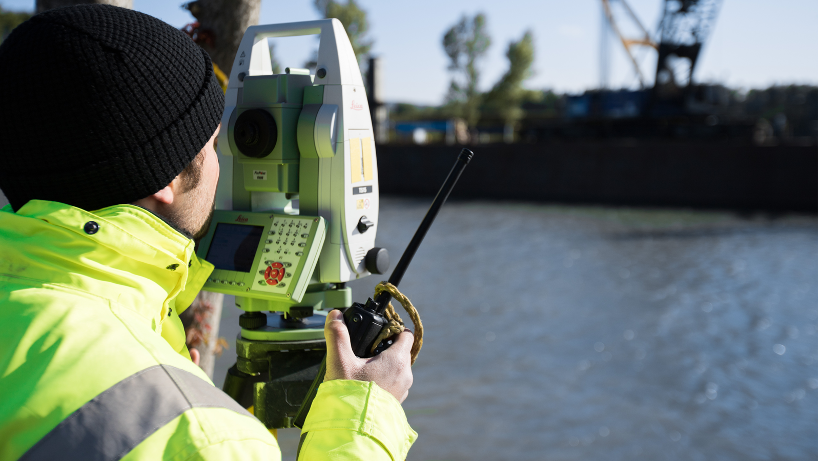 Mitarbeiter von Bogensberger Vermessung mit gelber Jacke und schwarzer Mütze bedient eine Totalstation am Ufer; im Hintergrund Wasser und ein Kran.