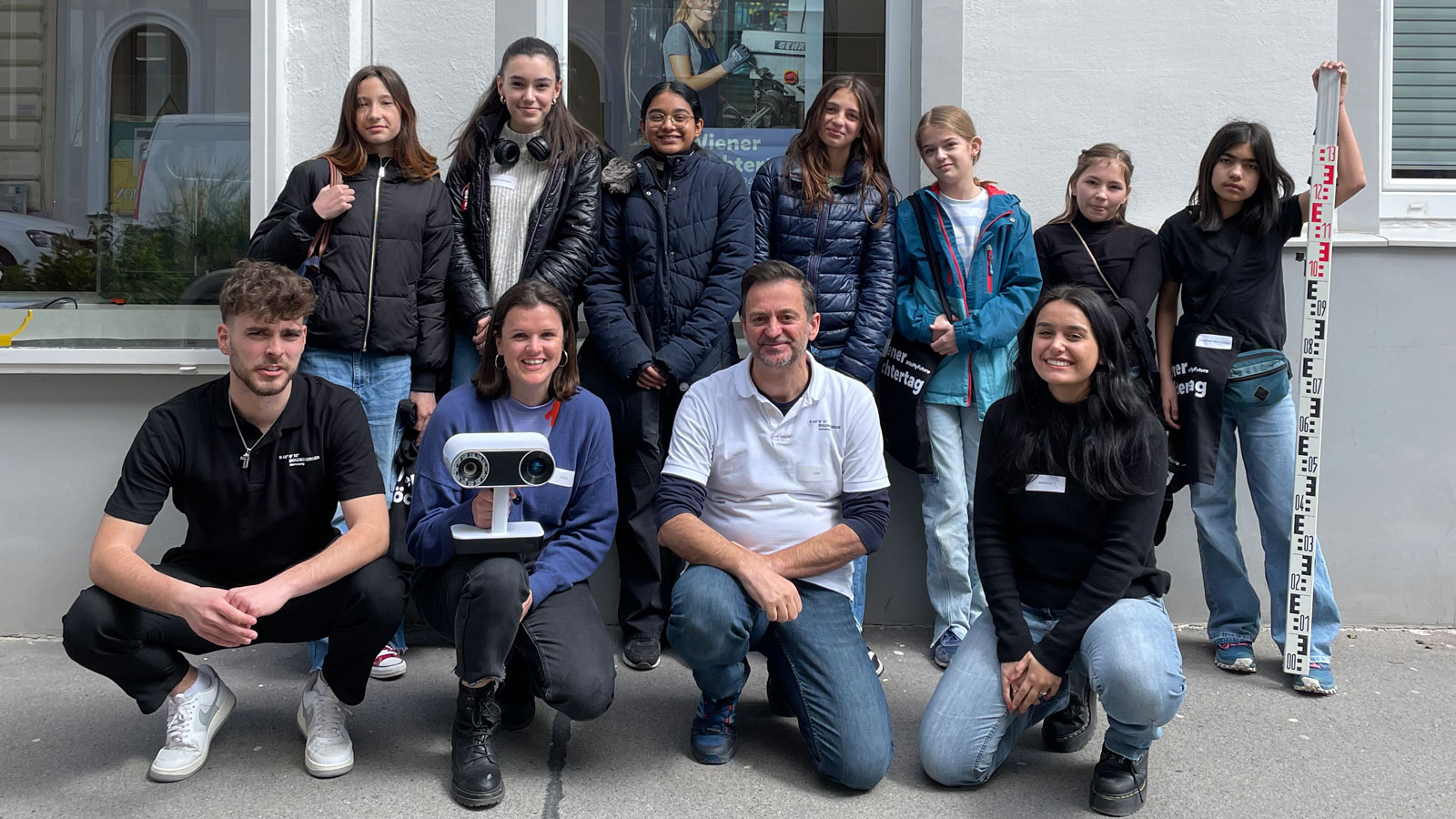 Gruppe junger Menschen posiert vor einer weißen Gebäudefassade; vorne knien vier Personen, darunter Beamer auf Stativ, rechts eine Messlatte sichtbar.