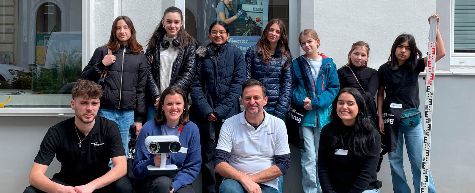 Gruppe junger Menschen und zwei Erwachsene posiert vor einer weißen Wand; vorne sitzt ein Mann im T‑Shirt, daneben ein Beamer, rechts hält eine Person eine Messlatte hoch.