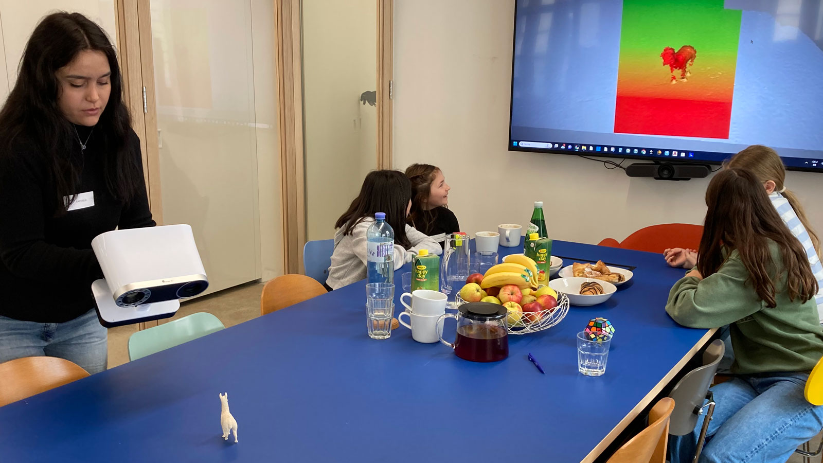 Jugendliche um einen blauen Tisch in einem Seminarraum; eine Person hält ein weißes Präsentationsgerät. Bildschirm zeigt Herzmotiv; Tisch mit Obst und Snacks.