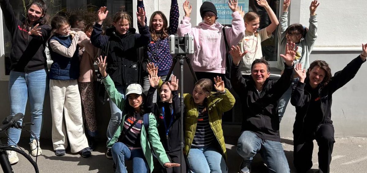 Eine Gruppe von Kindern und Erwachsenen beim Wiener Töchtertag 2026 hebt die Hände zum Gruppenfoto vor einer weißen Fassade mit Fenstern.